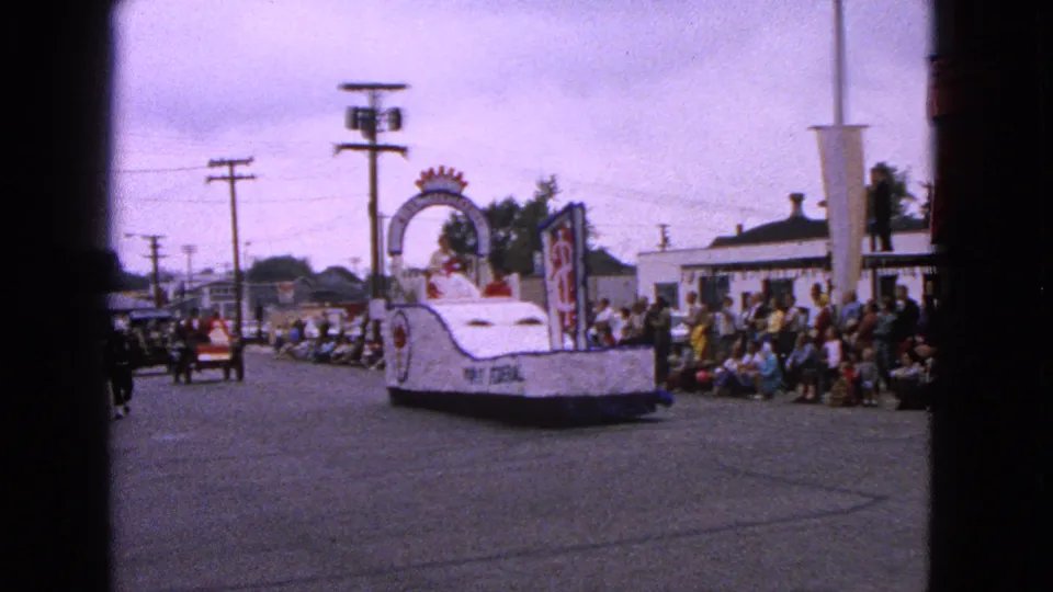 A white parade float moves through Barrington, Illinois, with curbside spectators, parked cars, utility poles, and a following vehicle visible.