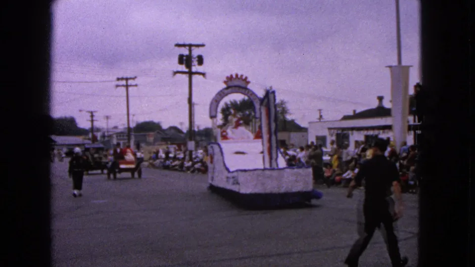 The rear of a white parade float continues along the Barrington street, with spectators at the curb and pedestrians crossing in front of the camera.