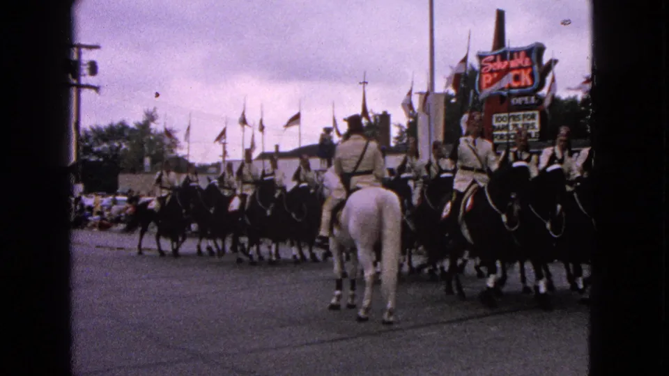 Uniformed horseback riders in the 1963 Barrington parade gather in close formation beside spectators and visible commercial signage.