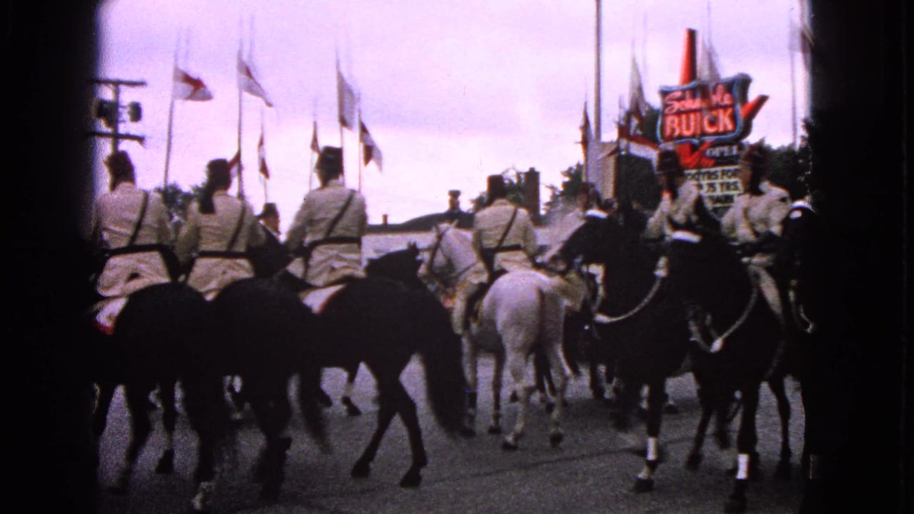 1963 Barrington, Illinois home-movie frame showing uniformed horseback riders moving past flags and a Schwab's Buick sign.