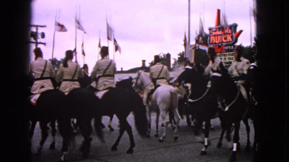 1963 Barrington, Illinois home-movie frame showing uniformed horseback riders moving past flags and a Schwab's Buick sign.