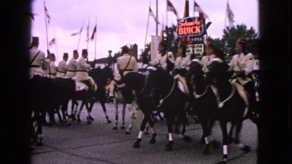 Uniformed horseback riders in Barrington, Illinois, seen from behind and side as they pass flags and period business signage.