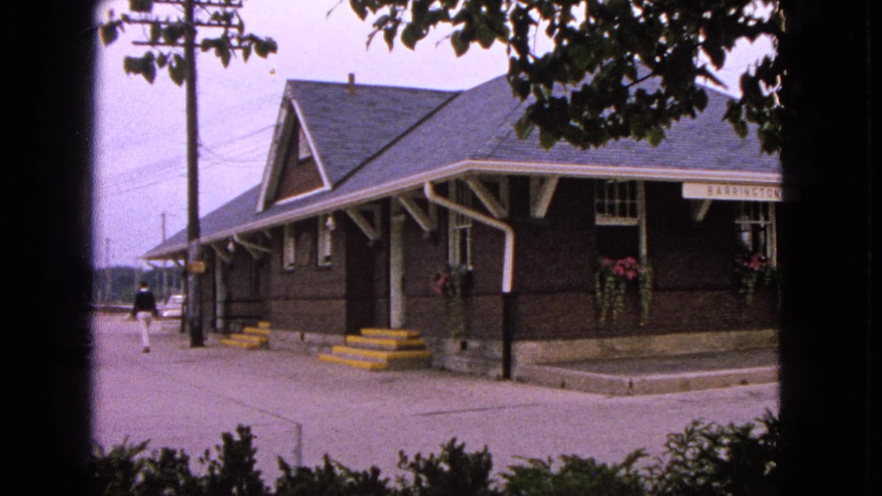 Barrington, Illinois train station exterior in 1963 home-movie footage, with yellow steps, hanging baskets, and a person on the platform.