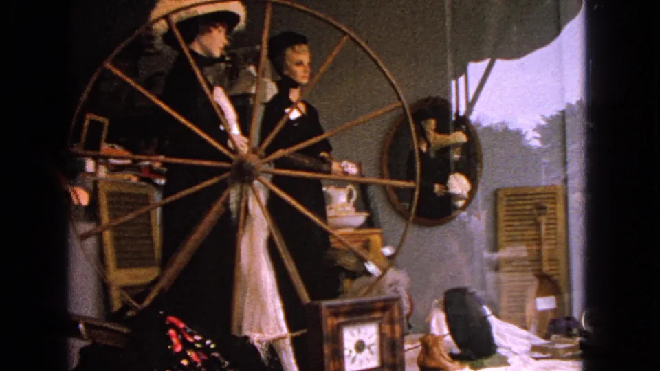 1963 home-movie view of mannequins in a Barrington display, with a large wooden spinning wheel, mirror, shutters, and small furnishings.