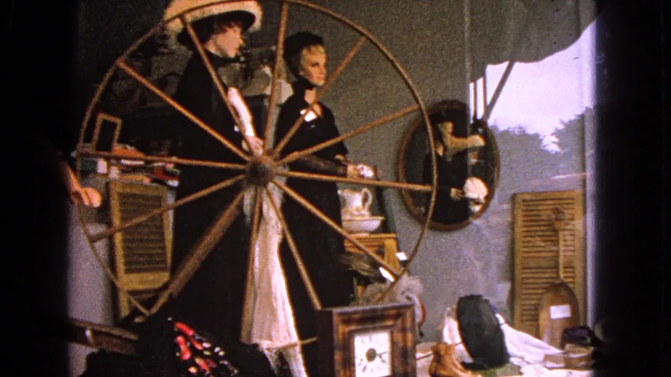 Barrington 1963 home-movie frame showing a storefront-style mannequin display with a wooden spinning wheel, shutters, mirror, and decorative objects.