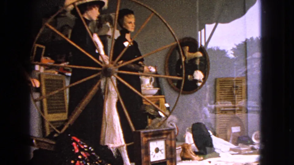 A 1963 Barrington display scene with mannequins, a wooden spinning wheel, an oval mirror, shutters, and window light at the edge of the frame.