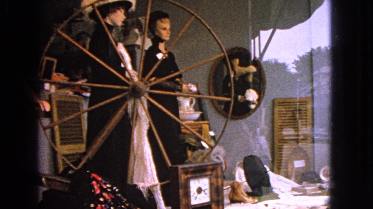 Shop window display with two mannequins behind a large wooden spinning wheel.