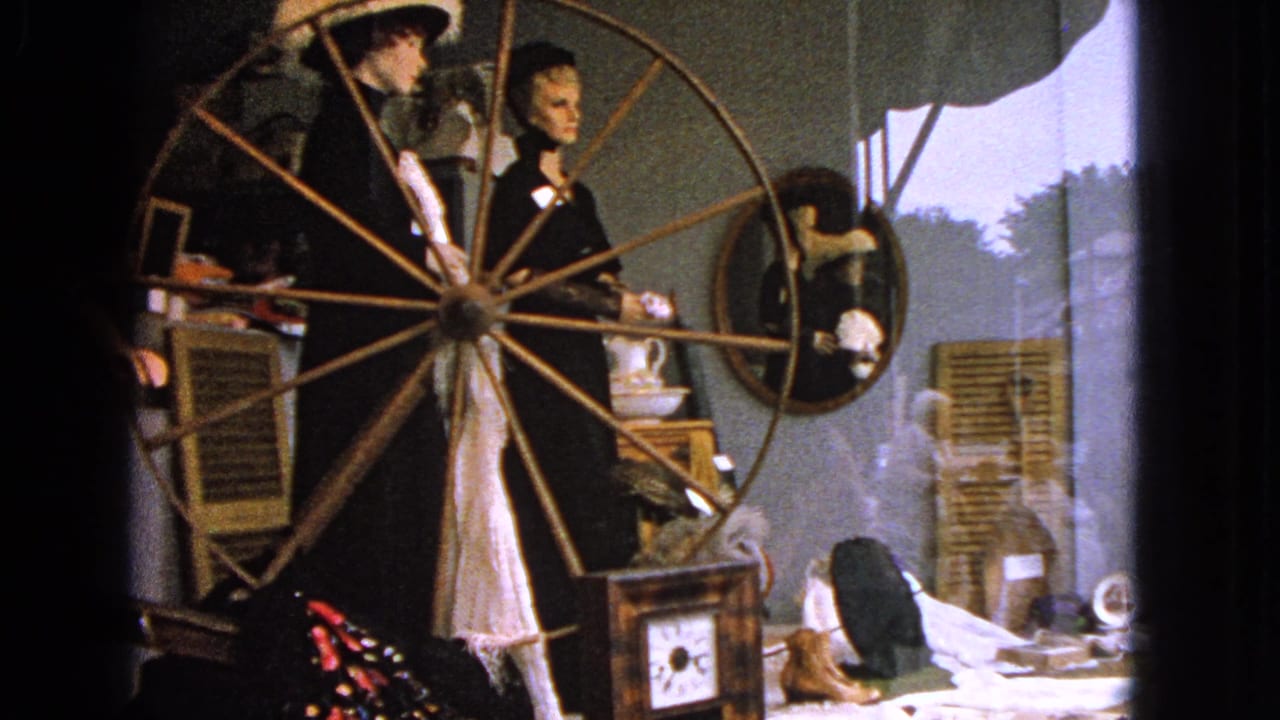 Mannequins, spinning wheel, mirror, and clock in a reflective shop window.