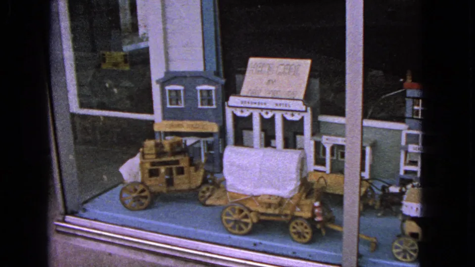1963 Barrington, Illinois home-movie frame showing a wooden Western town display with a covered wagon and storefronts behind glass.