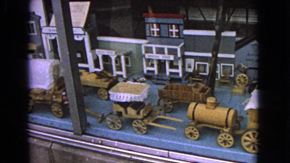 A closer 1963 home-movie view of the Barrington window display shows miniature storefronts, a small wagon, barrels, and wooden wheels.
