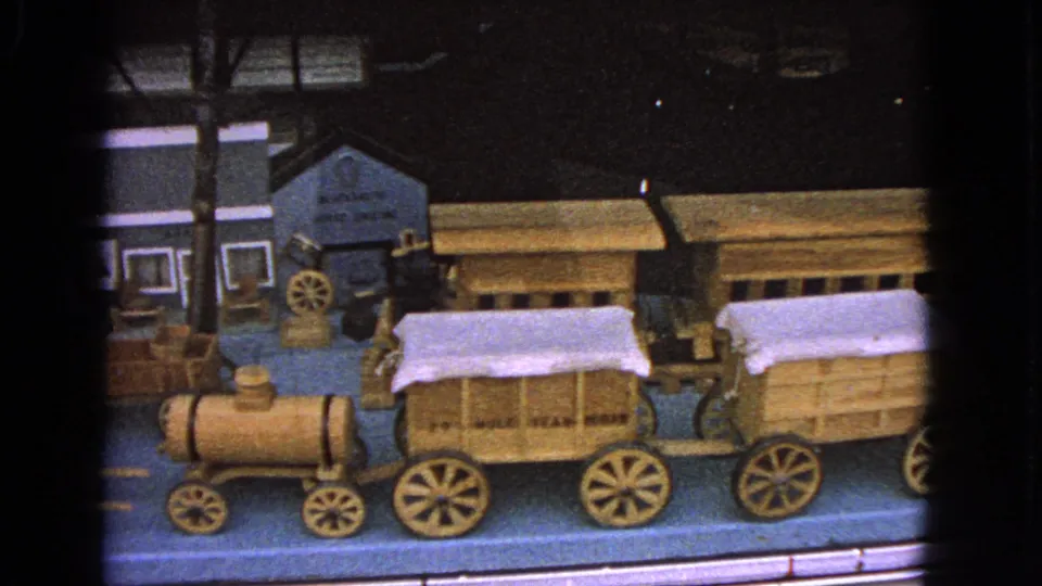 The final frame holds on the model display, with wooden covered wagons and a small engine passing across the foreground of shop facades.