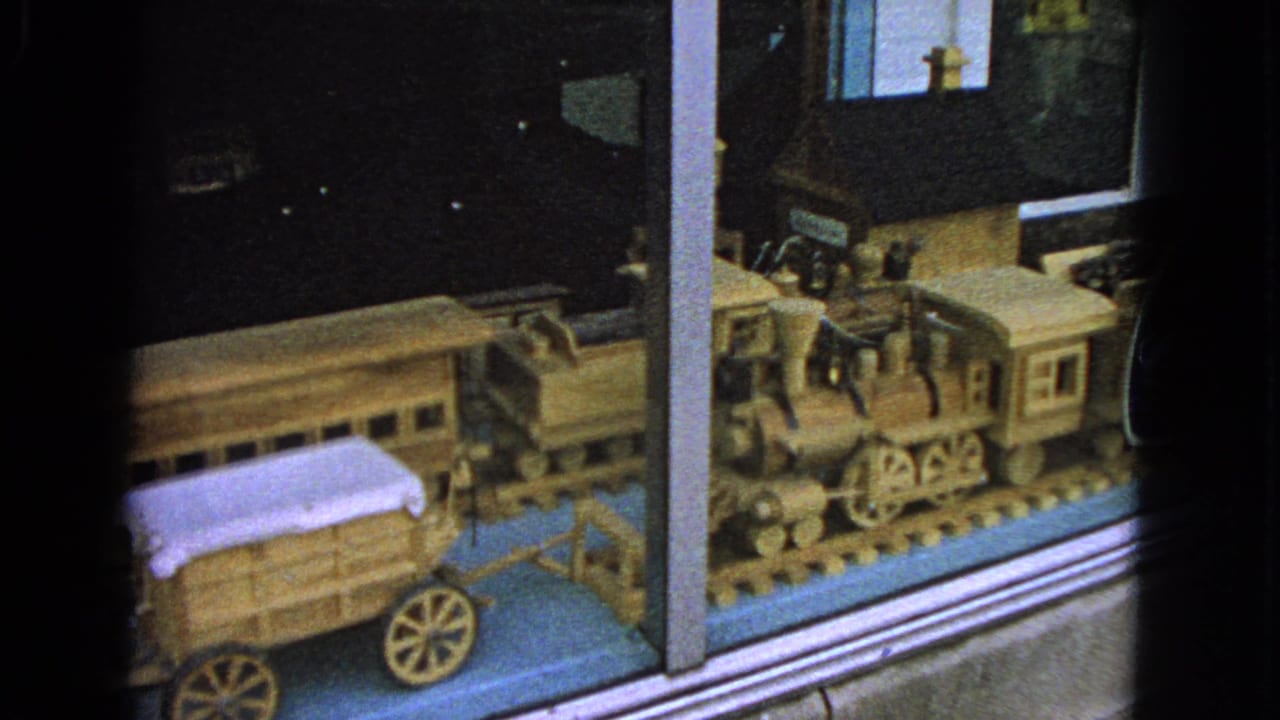 1963 Barrington, Illinois 8mm frame showing a wooden toy train set and covered wagon behind reflective glass.