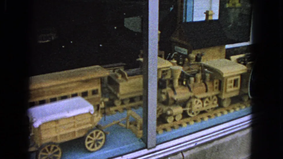 1963 Barrington, Illinois 8mm frame showing a wooden toy train set and covered wagon behind reflective glass.