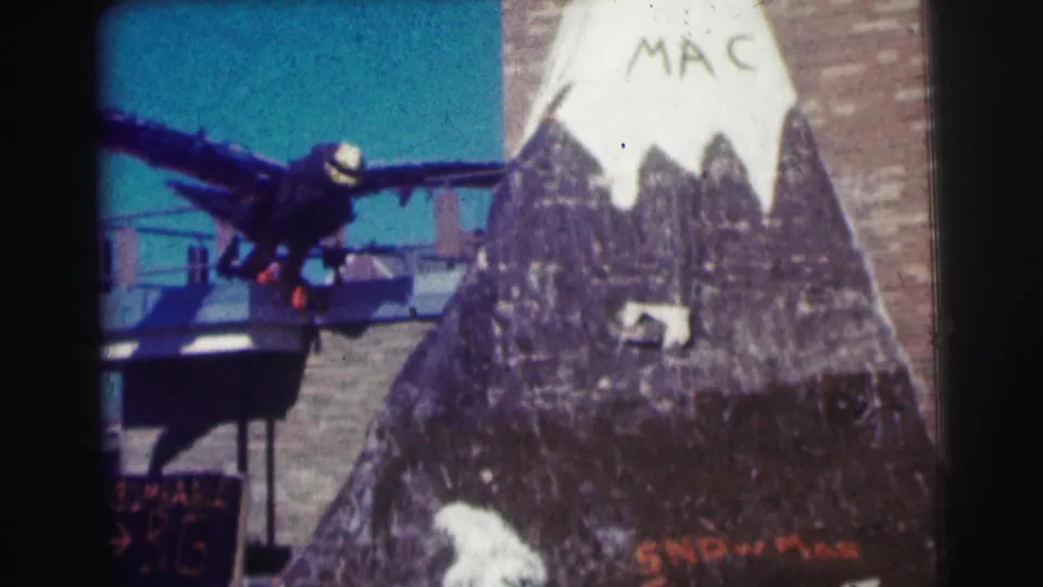 1963 Bowling Green University home-movie frame showing a dark winged bird figure near a painted mountain display marked MAC.