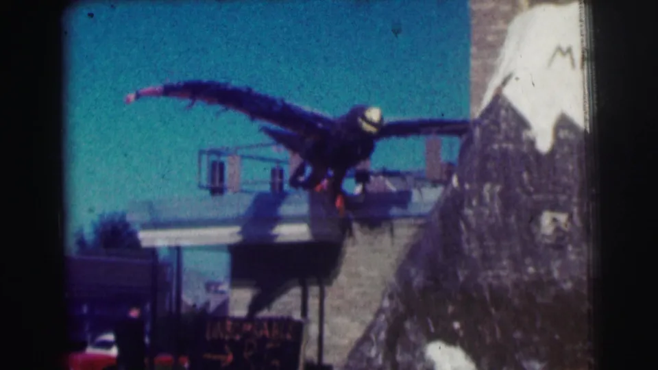 1963 8mm view at Bowling Green University showing a fake eagle prop perched over a building, with a sign and red car below.
