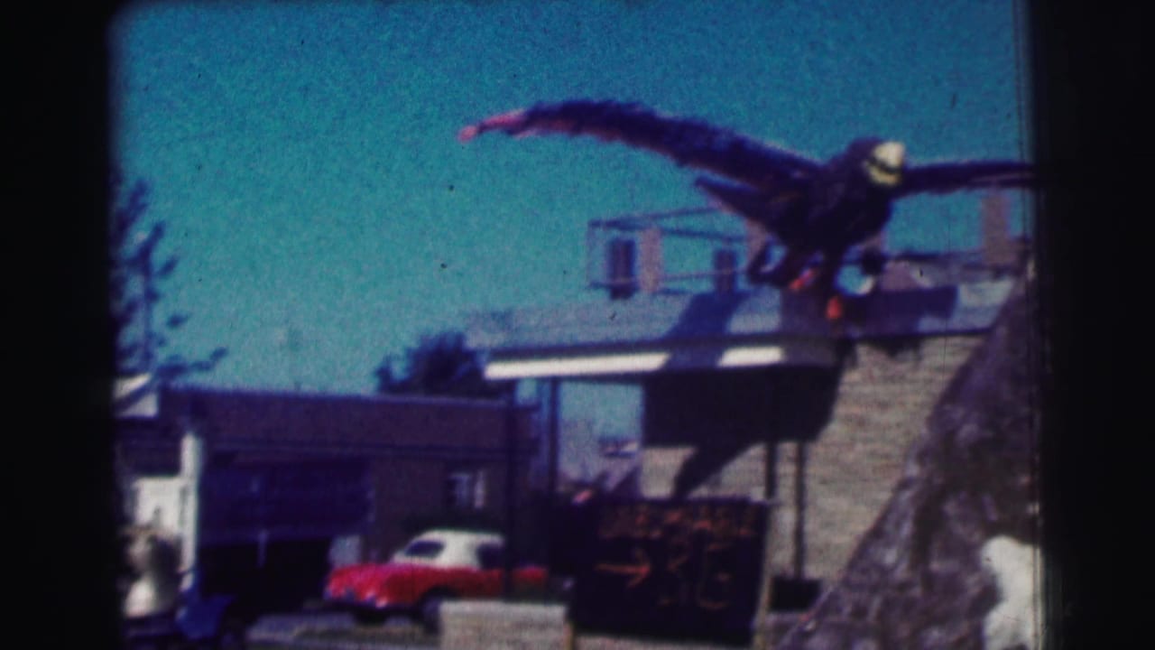 1963 home-movie frame at Bowling Green University with a large eagle prop on a building and a red car parked below.