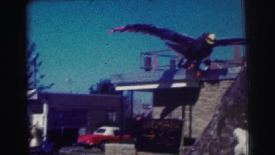1963 home-movie frame at Bowling Green University with a large eagle prop on a building and a red car parked below.