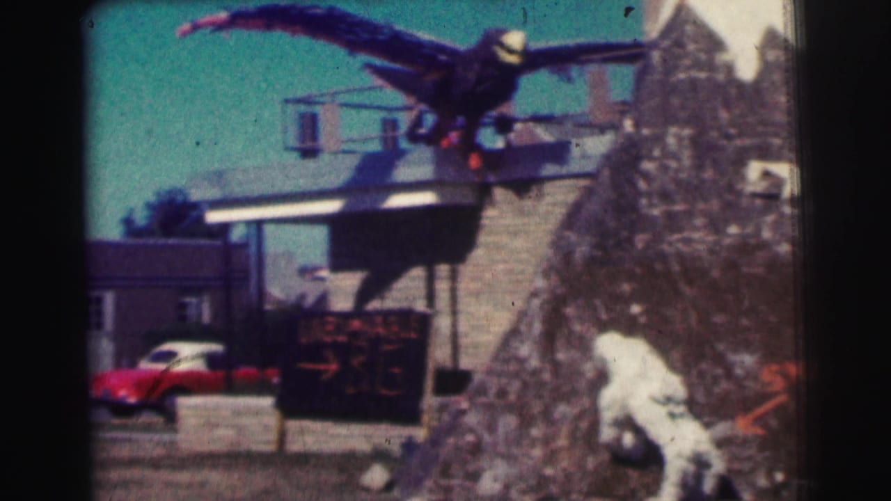 1963 8mm home-movie frame at Bowling Green University showing a large dark bird sculpture, small building, red car, and painted directional sign.