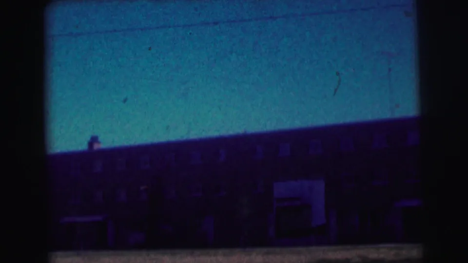 Blue-tinted 8mm frame showing a long brick university building with rows of windows and a shadowed entrance area.