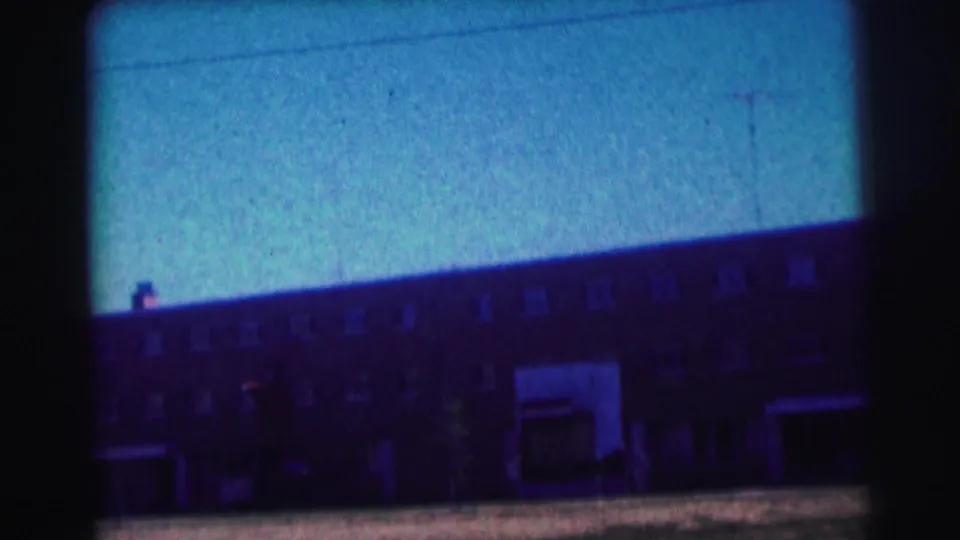 1963 campus exterior frame with a long brick building, repeated windows, open ground, and strong blue color cast from 8mm film.