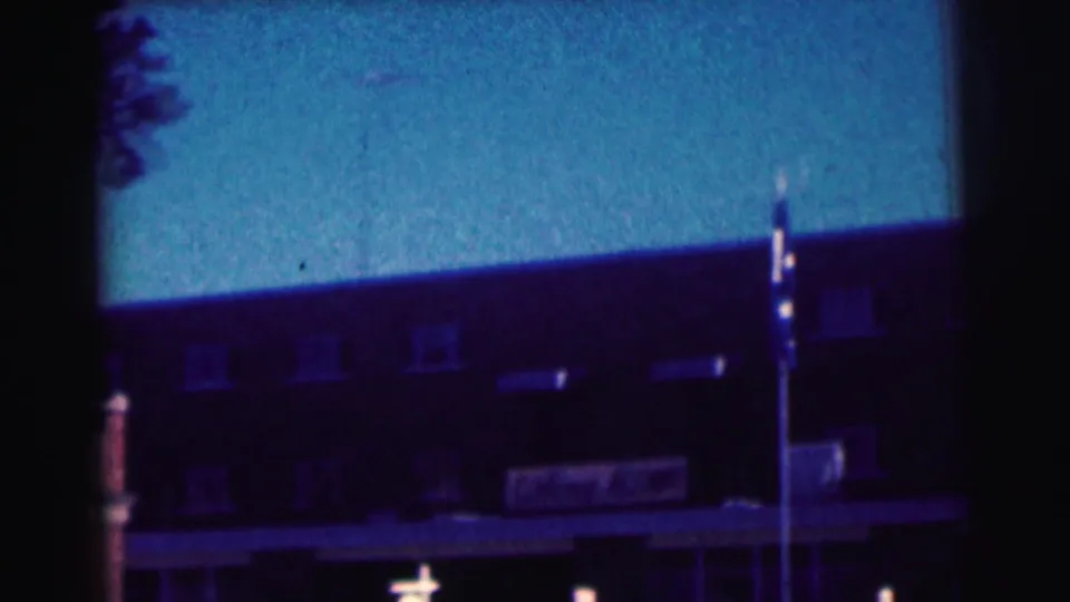 1963 8mm frame shows the university building facade with a flagpole and a dark tree edge in the foreground.