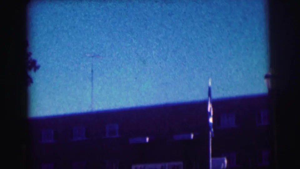 1963 8mm view at Bowling Green University showing a flagpole before a brick building with rooftop antennas and blue sky.
