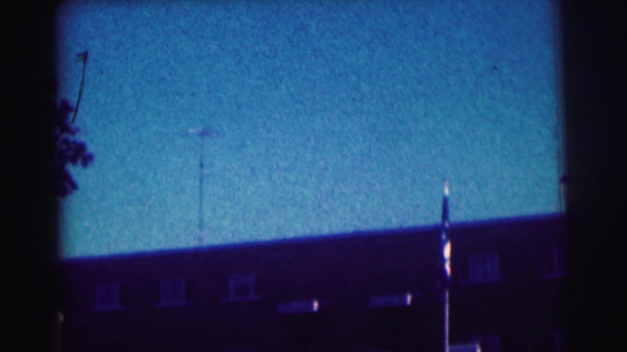 Flagpole, brick building, antennas, and blue sky framed by dark film edges.