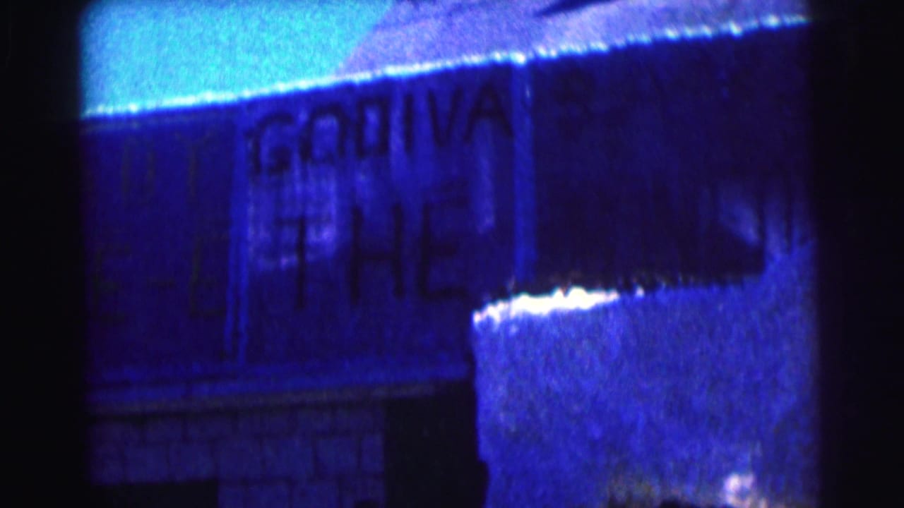 1963 8mm frame at Bowling Green University showing a close, soft-focus view of Godiva lettering on a sunlit building wall.