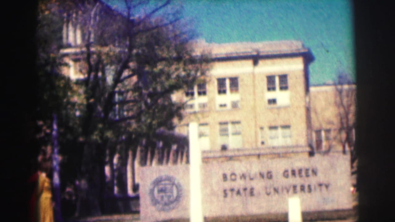 1963 8mm view of a Bowling Green State University stone sign before a brick academic building, with trees and blue sky.