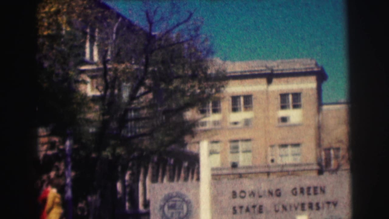 Bowling Green State University sign and brick academic building, partly obscured by trees, with a person visible near the frame edge.