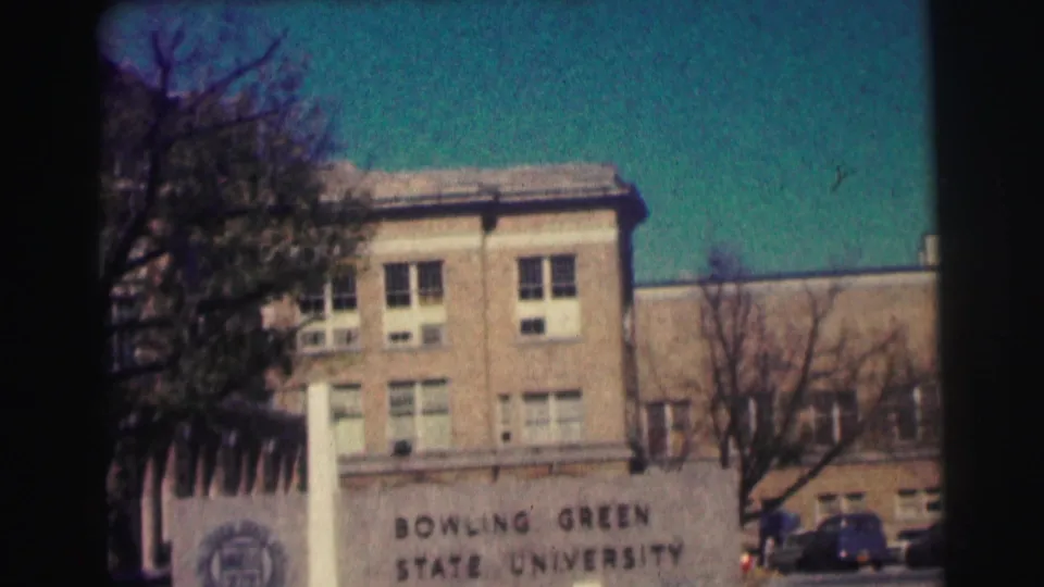 1963 campus exterior showing the Bowling Green State University sign, brick building, bare trees, and parked mid-century cars.