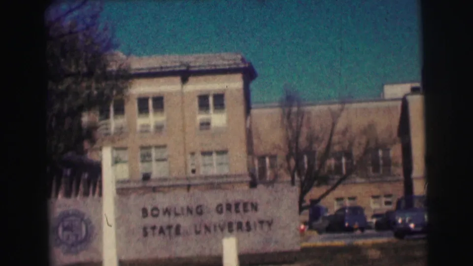 End frame of the Bowling Green State University sign before a brick campus building, with trees and parked cars visible.