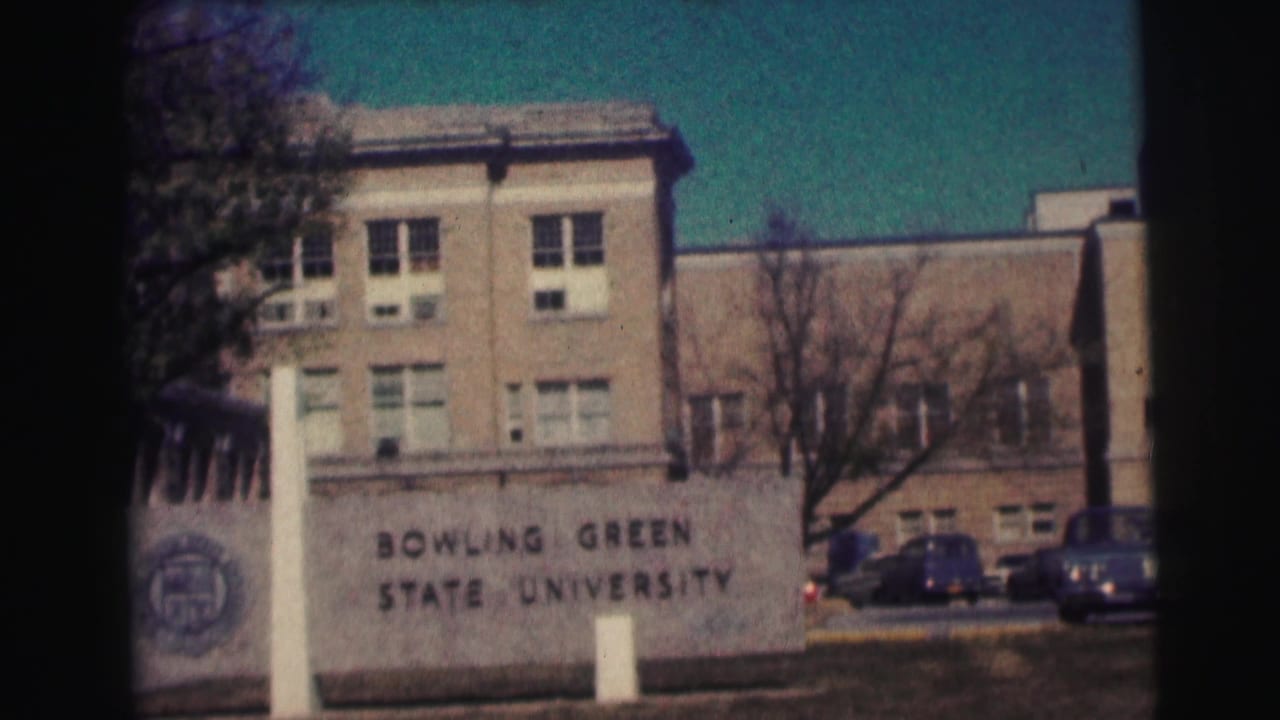 1963 8mm frame showing the Bowling Green State University sign with campus buildings, leafless trees, and parked cars behind it.