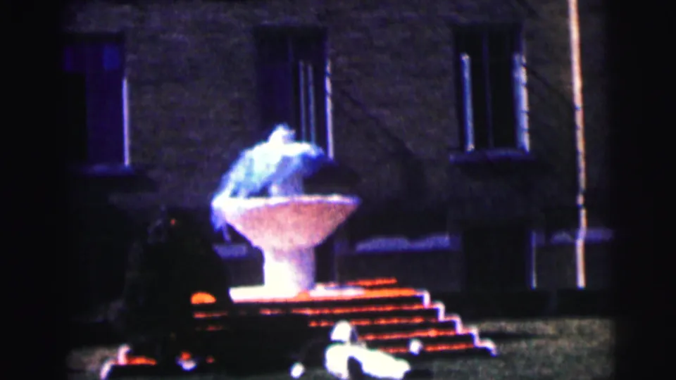 Night campus frame showing the illuminated fountain, steps, brick building, and a partially visible man near the foreground.