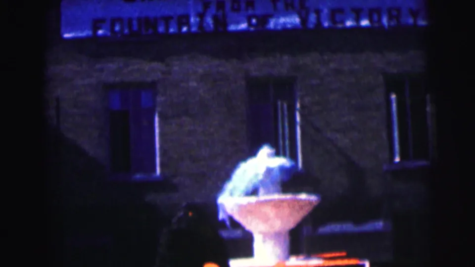 Nighttime 8mm frame of the Fountain of Victory area with a partial overhead sign and brick building behind it.