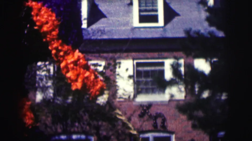 1963 campus travel frame showing a residential-style exterior with windows, roofline, shadows, and orange autumn foliage.