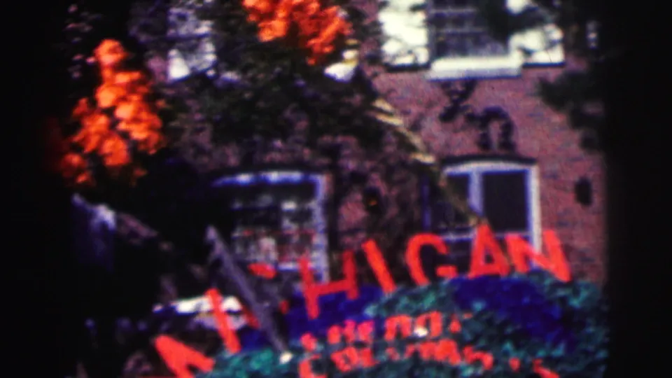 Home-movie frame from the Bowling Green University clip showing autumn foliage, a building exterior, and a red Michigan sign.