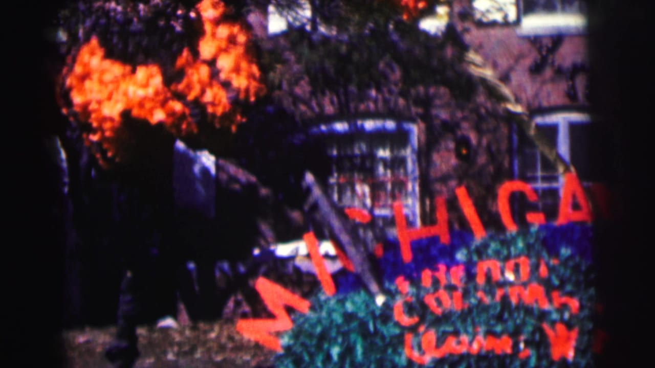 1963 8mm frame at Bowling Green University showing a red MICHIGAN garden sign, shrubs, orange flowers, and brick campus building details.