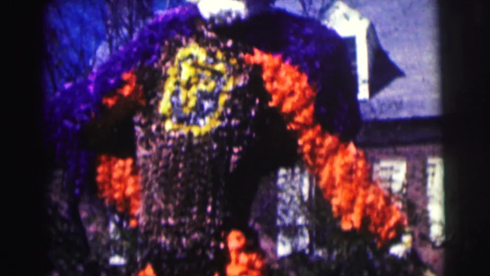 The frame shifts to a figure-like floral arrangement with orange, purple, yellow, and dark plantings against a brick building backdrop.