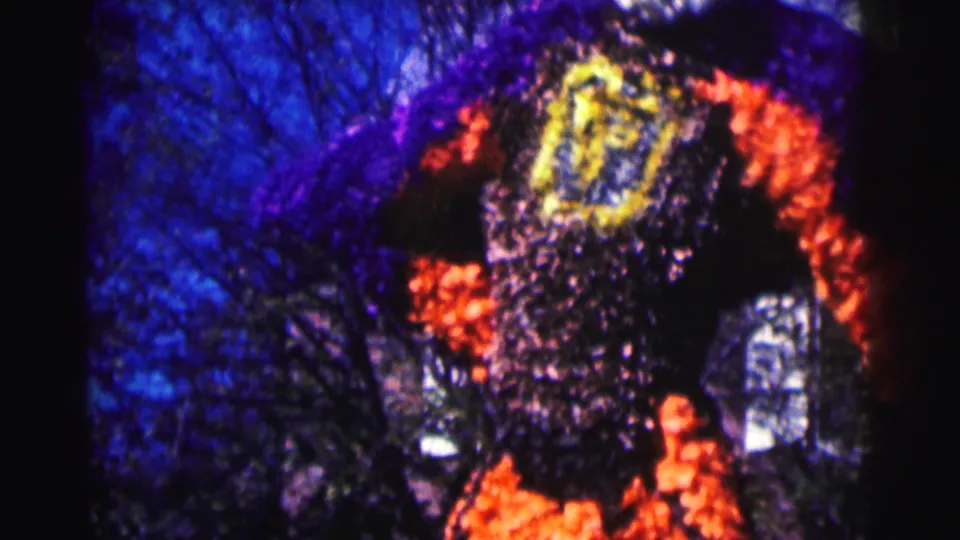 1963 8mm frame showing the torso of a large purple and orange floral mascot or float with a yellow emblem on its chest.