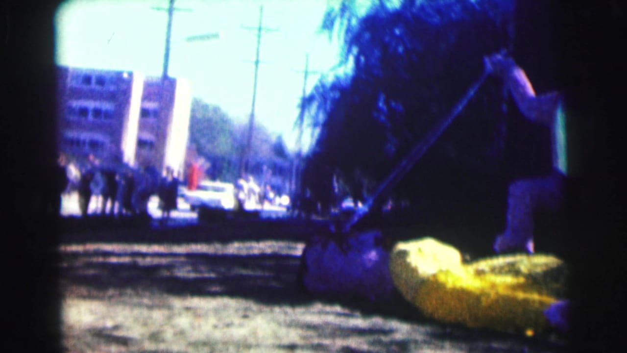 Street-side float scene with yellow prop, spear, trees, cars, utility poles, and apartment building.
