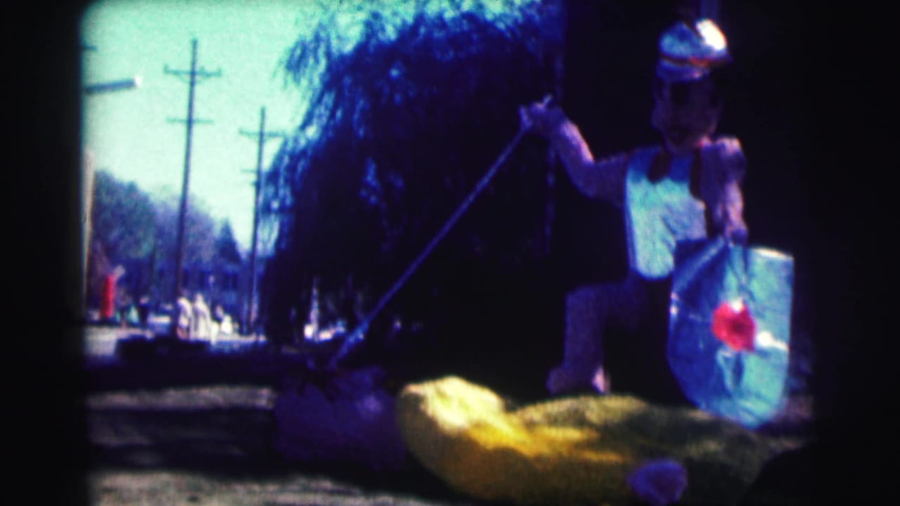 1963 Bowling Green University frame of a helmeted costumed figure holding a spear and shield beside a yellow float prop on a street.