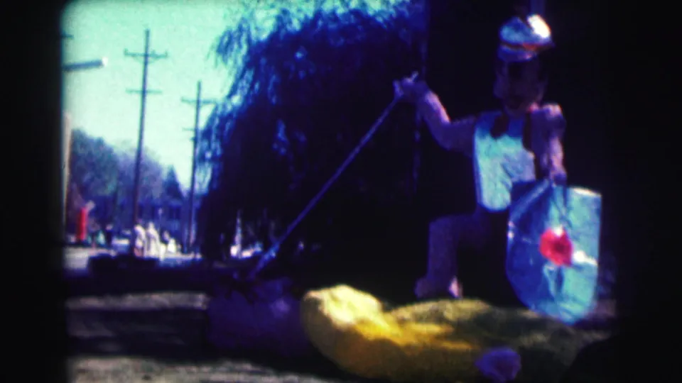 1963 Bowling Green University frame of a helmeted costumed figure holding a spear and shield beside a yellow float prop on a street.