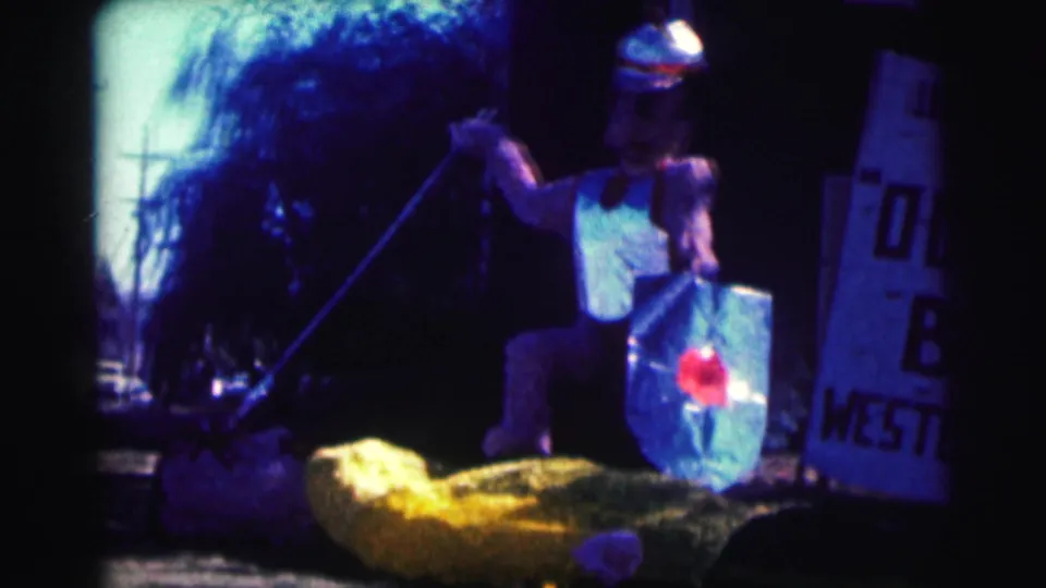 1963 Bowling Green University home-movie frame with a costumed spear-and-shield figure beside a yellow prop and partial hand-lettered sign.