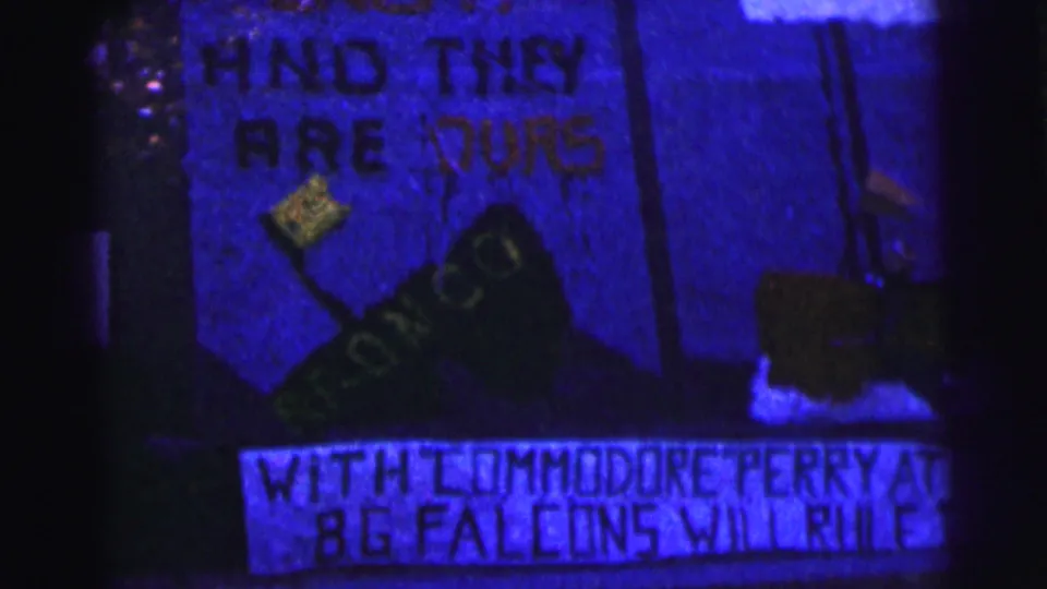 Blue-tinted 1963 Bowling Green University frame showing a handmade sign with partial text, a shoe graphic, and a lower banner about Commodore Perry and B.G. Falcons.