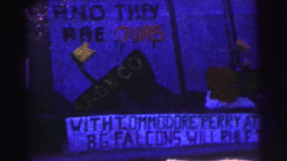 Close 1963 campus protest frame showing partly readable hand-painted wording and a dark exterior wall.