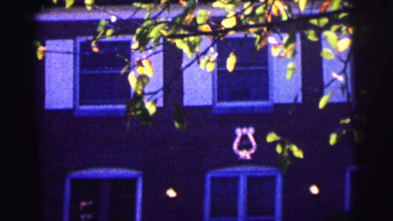 1963 Bowling Green University home-movie frame showing a purple-lit brick housing facade and tree branches at night.