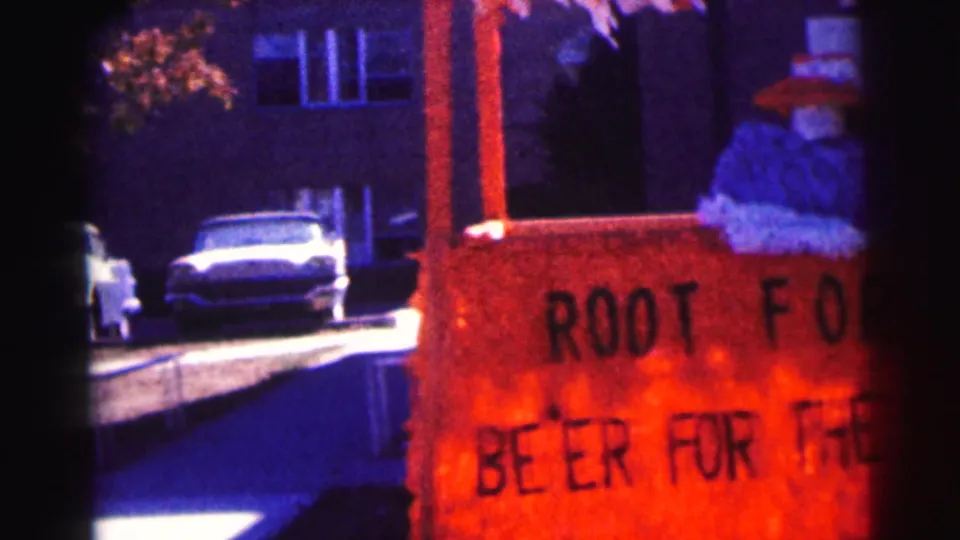 1963 campus street view with parked cars and a red game-support sign partially visible in front of brick buildings.