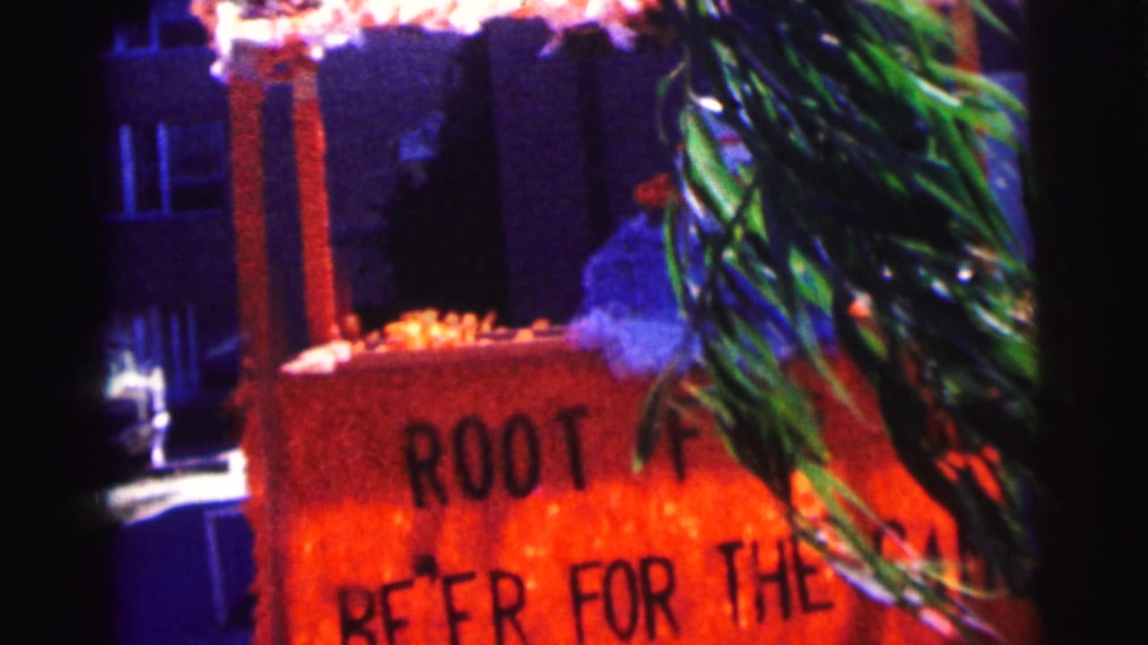 1963 Bowling Green University home-movie frame showing a handmade orange root beer sign with greenery in front and building windows behind.