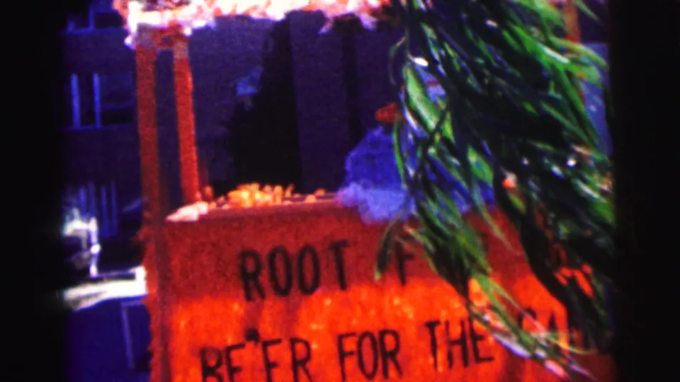 1963 Bowling Green University home-movie frame showing a handmade orange root beer sign with greenery in front and building windows behind.